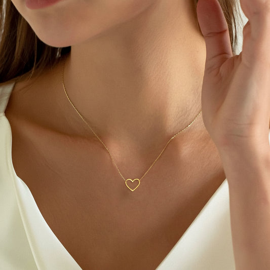 A close-up of a woman wearing a delicate gold necklace with a small open heart pendant, styled with a white top.